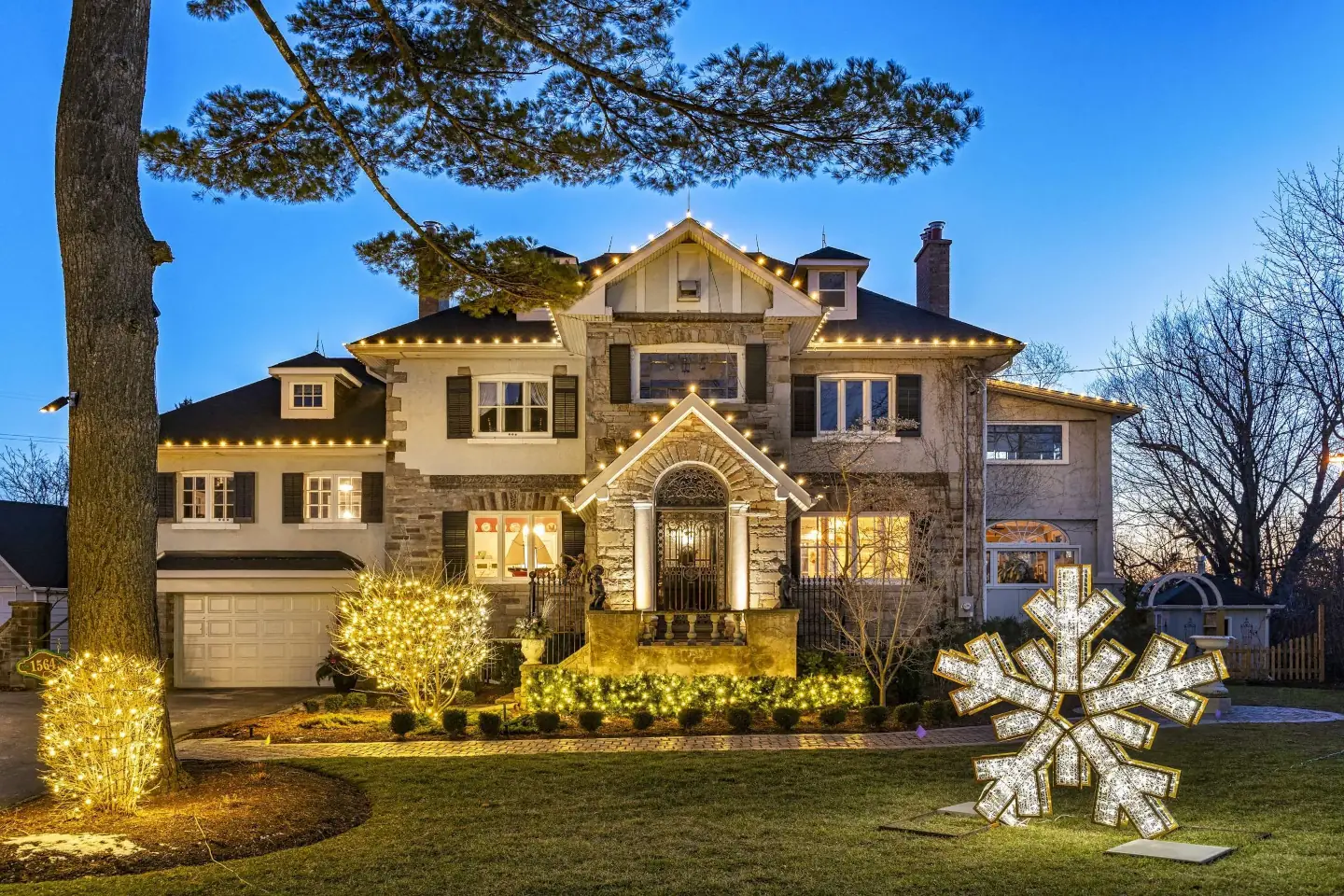 Luxury home decorated with warm white Christmas lights on the roofline, trees, and landscaping, featuring an illuminated snowflake display.