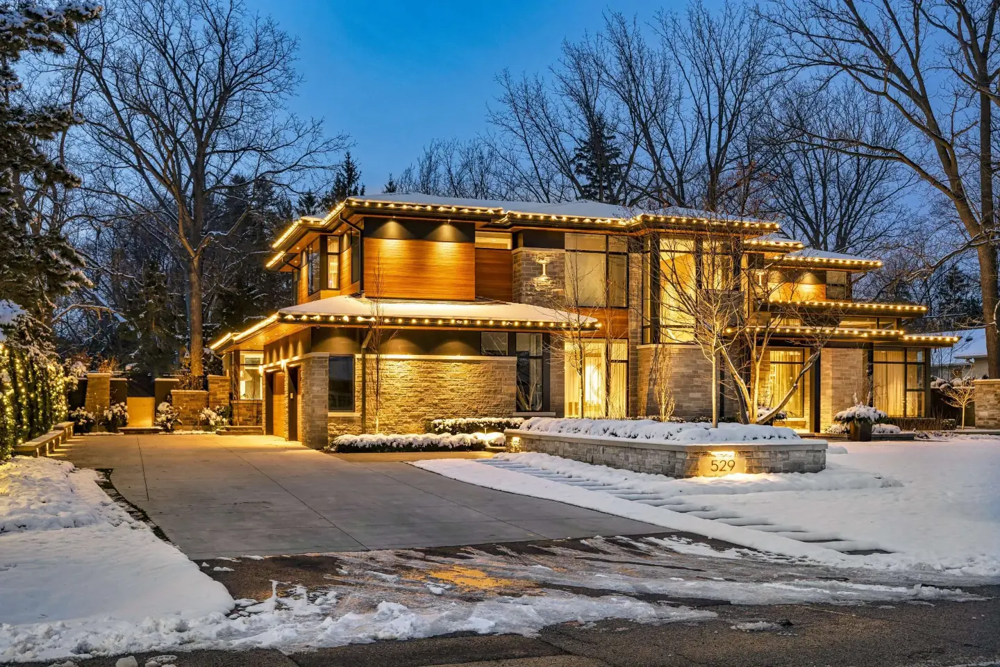 Modern home decorated with warm white Christmas lights along the roofline and landscaping, surrounded by snow.