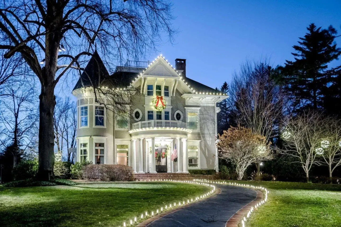 Victorian-style home decorated with warm white Christmas lights on the roofline, walkway, and trees, featuring a festive wreath.